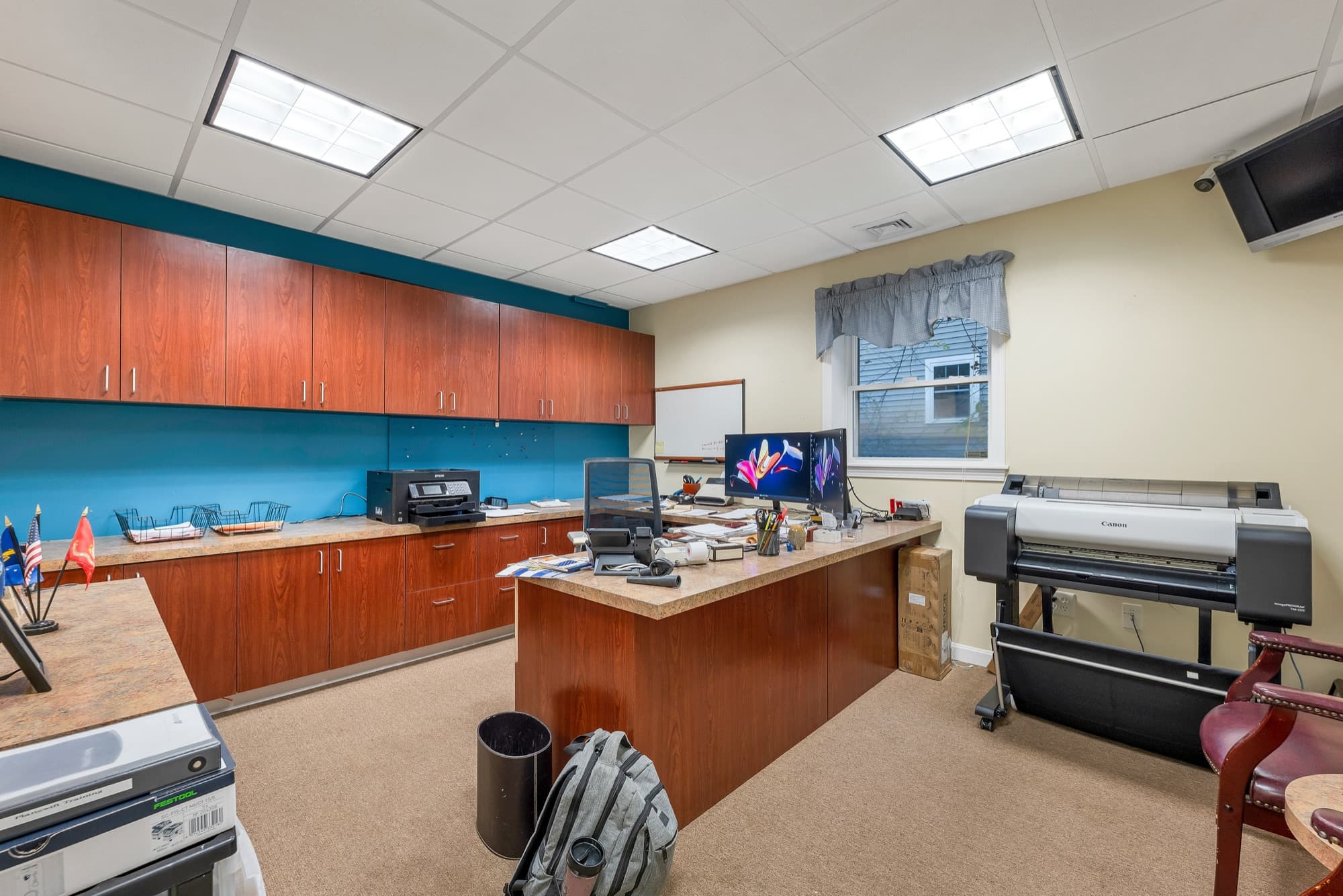 An office with cherry upper cabinets along a teal wall, a desk, and a wide-format printer