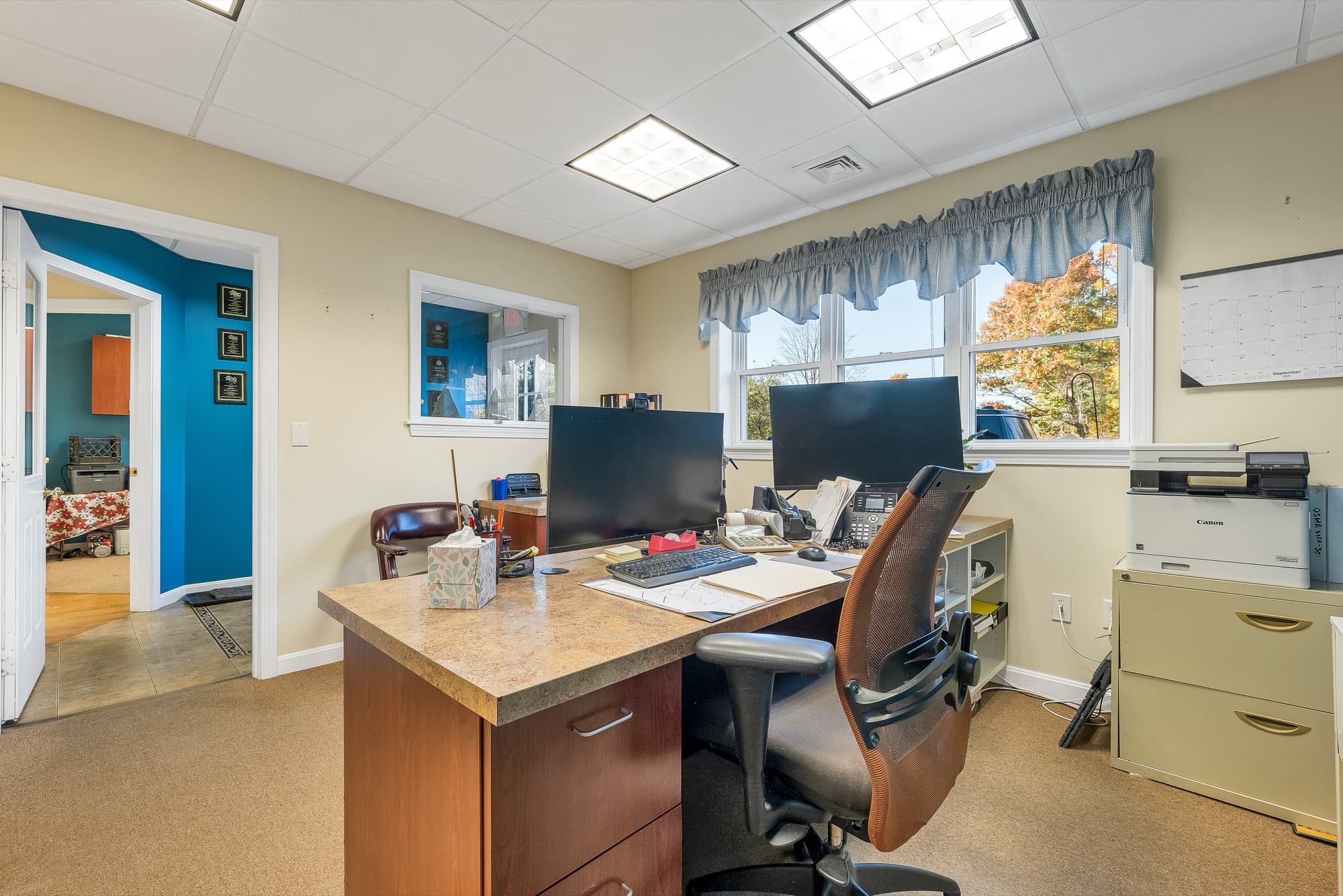 An office with a window, a desk with 2 monitors, and a printer, looking into the hall